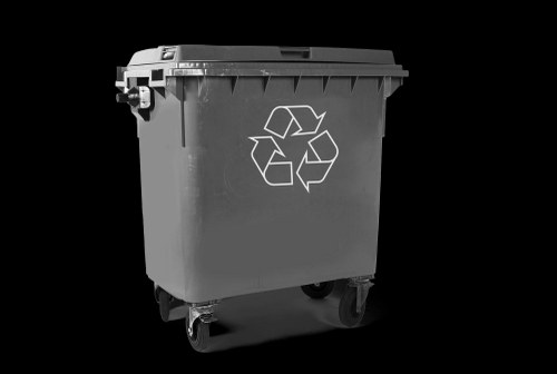 A large black wheeled rubbish bin made of durable plastic with a hinged lid and a prominent white recycling symbol on its front. The bin is positioned outdoors on a paved surface, likely in a driveway or storage area, with a smooth finish and slightly textured surface. The lid is closed, and the bin appears to be in good condition, designed for waste collection and recycling purposes. In the background, a subtle hint of an outdoor environment may be present, suggesting that the bin is ready for waste disposal or collection services. As part of commercial waste management, this bin is associated with local rubbish removal services such as those offered by Commercial Waste Kingston upon Thames and is suitable for use in Kingston upon Thames or surrounding areas within postcode regions. The focus on the bin’s structure, materials, and placement reflects typical waste collection infrastructure in urban and suburban environments, supporting environmentally responsible waste disposal in the community.
