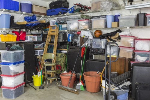 The image shows a cluttered storage area filled with various household and gardening items, including multiple clear plastic storage boxes stacked on shelves, some with coloured lids, along with cardboard boxes, a black toolbox, and a blue suitcase placed on the upper shelf. In the foreground, there are two plastic buckets—one terracotta-colored and the other brown—containing cleaning tools like brooms and mops leaning against a metal trolly. Several wooden chairs are stacked or arranged near the buckets, and a small yellow bucket with a mop head is also visible. The environment appears to be a garage or utility room with concrete flooring and metal shelving units, indicative of a space used for storage and organisation. The overall scene is busy with various bulky and miscellaneous items that likely require removal or disposal, aligning with the services offered by Commercial Waste Kingston upon Thames in the local area. Natural lighting highlights the different textures and materials, including plastic, wood, and metal, emphasizing the cluttered state typical of a commercial or domestic waste collection context in Kingston upon Thames.