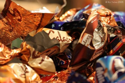 A close-up image of a pile of crumpled foil wrappers and candy packaging materials, featuring metallic silver, gold, and copper tones with some colorful printed designs and logos visible on the wrappers. The wrappers are overlapping each other, creating a textured and glossy surface that reflects light. The background shows more discarded wrappers, suggesting a collection of rubbish likely associated with sweet wrappers and snack packaging. The setting appears to be indoors, with the wrappers resting on a surface, possibly on a table or floor. This visual relates to waste collection and rubbish removal services, such as those provided by Commercial Waste Kingston upon Thames, especially in residential or commercial contexts within the Kingston area. The detailed description emphasizes the materials' textures and colours, aligning with environmental waste management and rubbish disposal activities in the local borough.