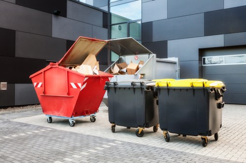 The image shows three large waste bins placed on a paved outdoor surface in front of a modern building with dark grey and black exterior panels. The first waste container, positioned on the left, is bright red with a partially open lid revealing cardboard boxes and paper waste inside. The surface of the red bin appears metallic with a smooth, painted finish and small caster wheels at the base for mobility. Next to it, there are two larger, closed black waste bins with yellow lids, also fitted with caster wheels for easy movement. The yellow lids are securely closed, indicating they are for waste collection. The backdrop features a contemporary structure with reflective glass and sleek panel cladding, suggesting a commercial or office environment in Kingston upon Thames. The scene is well-lit, with natural daylight highlighting the textures of the waste containers and the paved ground, providing a clear visual context related to rubbish management services provided by Commercial Waste Kingston upon Thames.