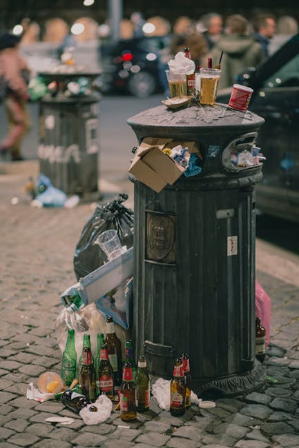 A black, cylindrical public bin situated on a cobblestone street with signs of wear and chipped paint. The bin's open top is cluttered with discarded paper cups, liquid containers, and various food wr