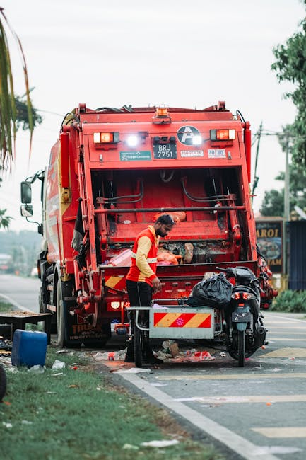 A narrow urban alleyway filled with a large, black plastic rubbish bag in the foreground, placed on a rough, uneven asphalt surface with minor debris scattered around. Behind the bag, there are additi